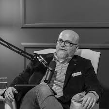 Dan Flowers, a man with a white beard in a smart suit, sits in an armchair with a microphone before him.